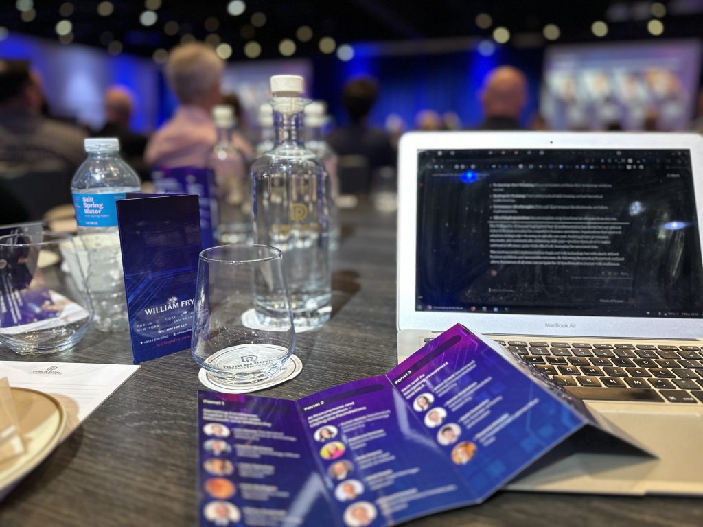 Close-up of a laptop and printed speaker agenda at a tech business conference, with attendees and stage in the background